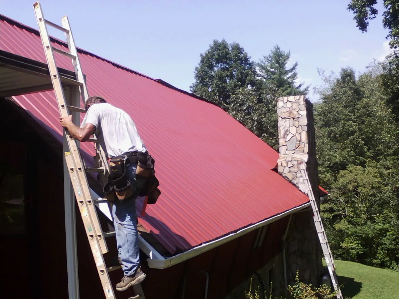 Roofer on a ladder beside a red metal roof and stone chimney — Soffit & Fascia Repair by Roofing Brothers in Holliston