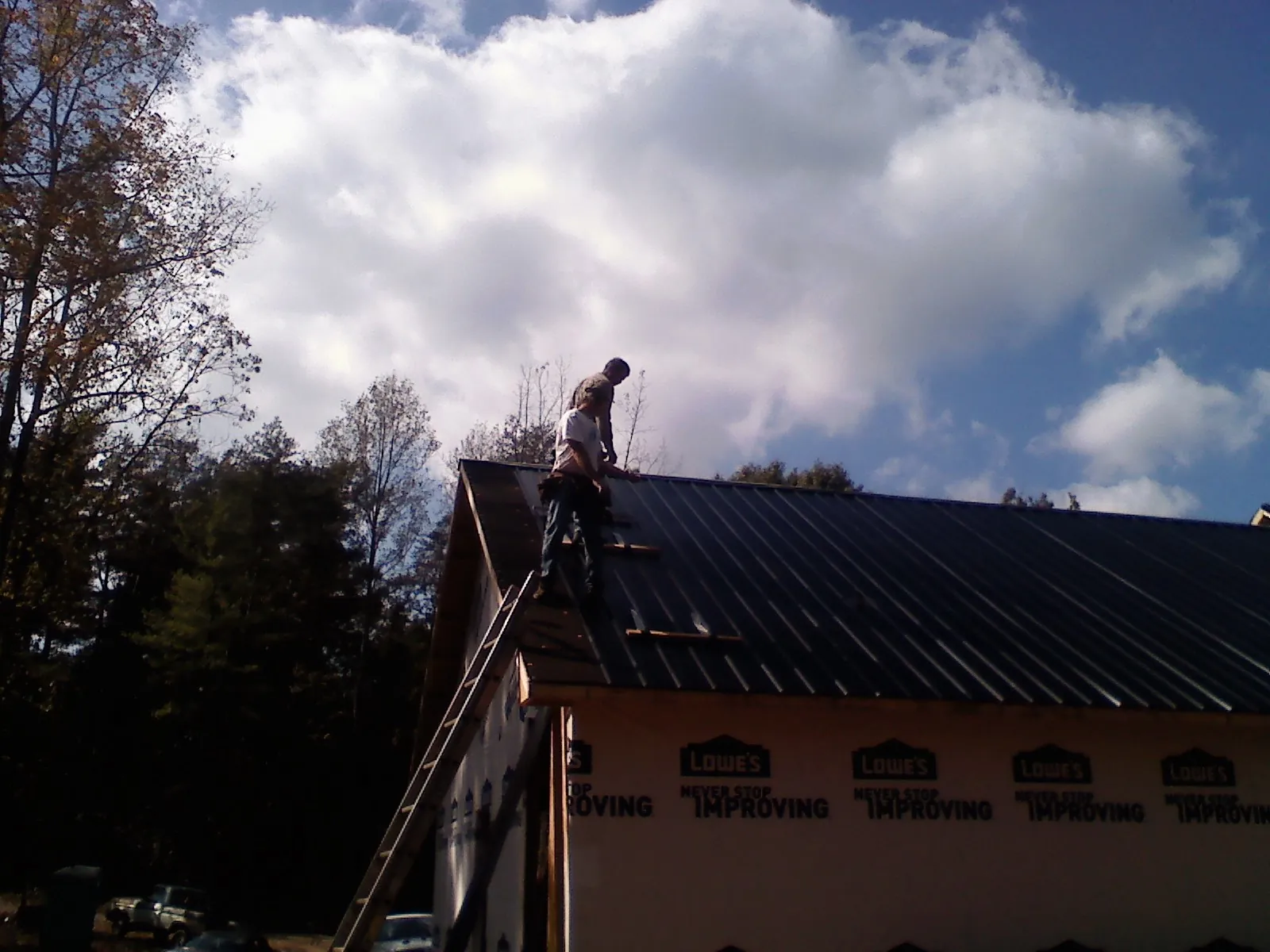 Roofer fastening dark metal panels high against a cloudy sky, from a Roof Inspection job in Holliston